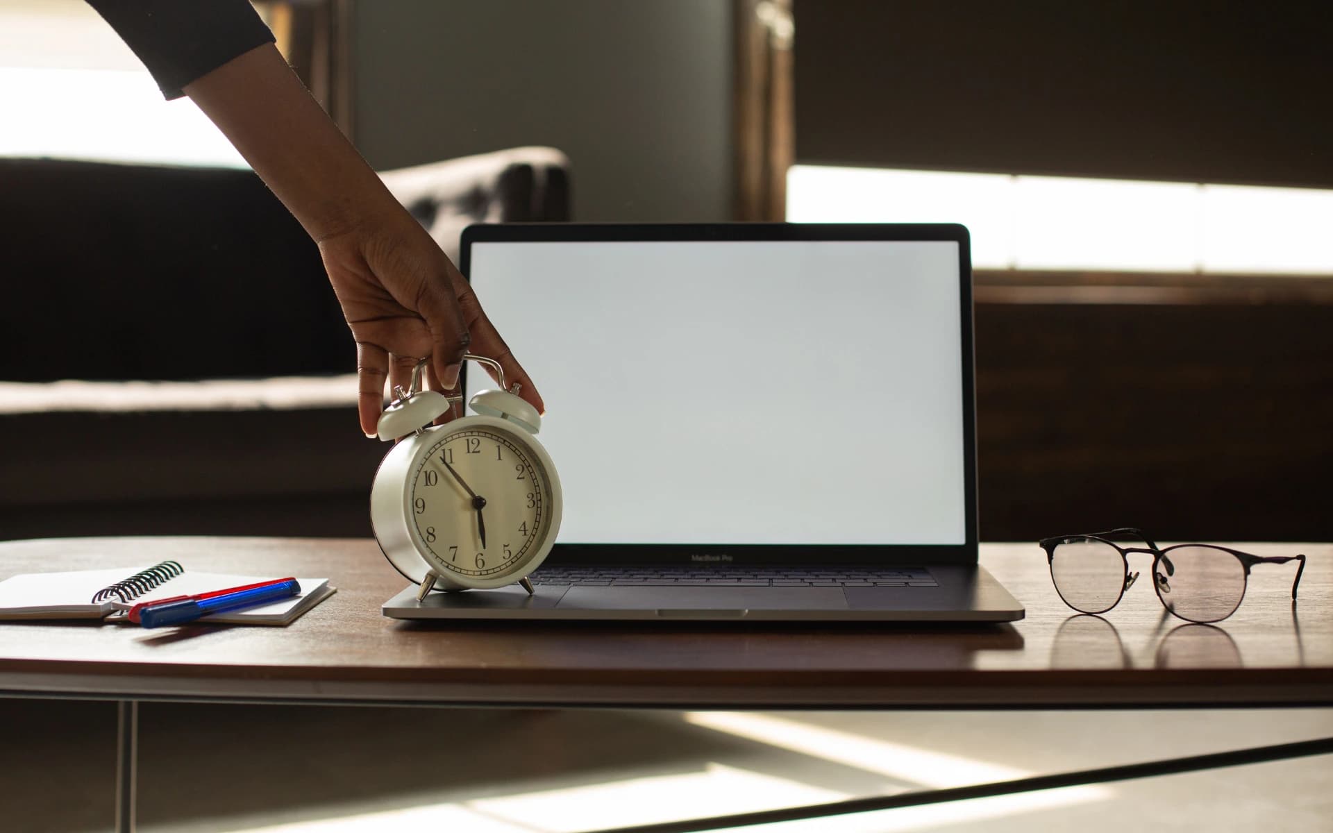 A hand reaching for an alarm clock standing beside an open laptop on a desk, early light coming through the blinds